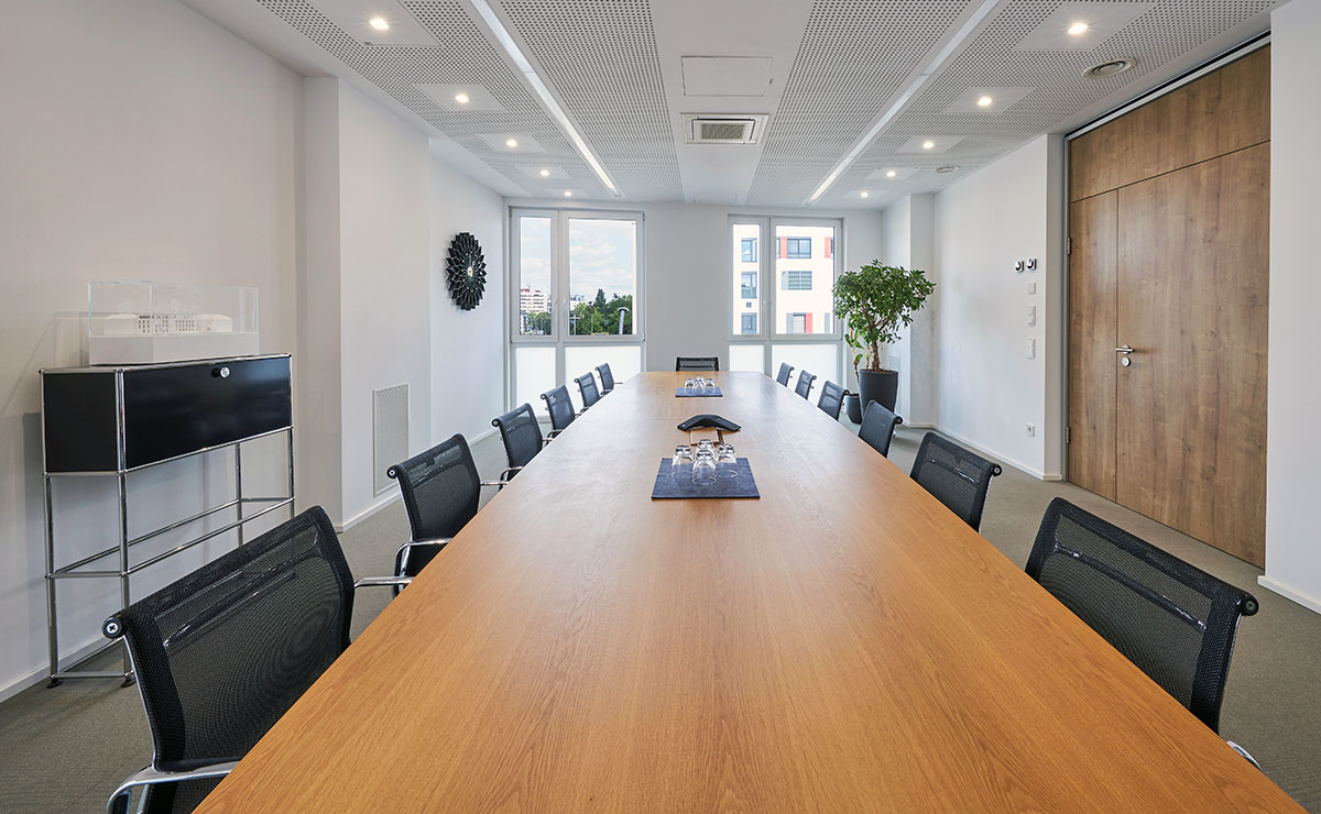 Moderner Konferenzraum mit langem Holztisch, mehreren B&uuml;rost&uuml;hlen, gro&szlig;en Fenstern und heller Beleuchtung.