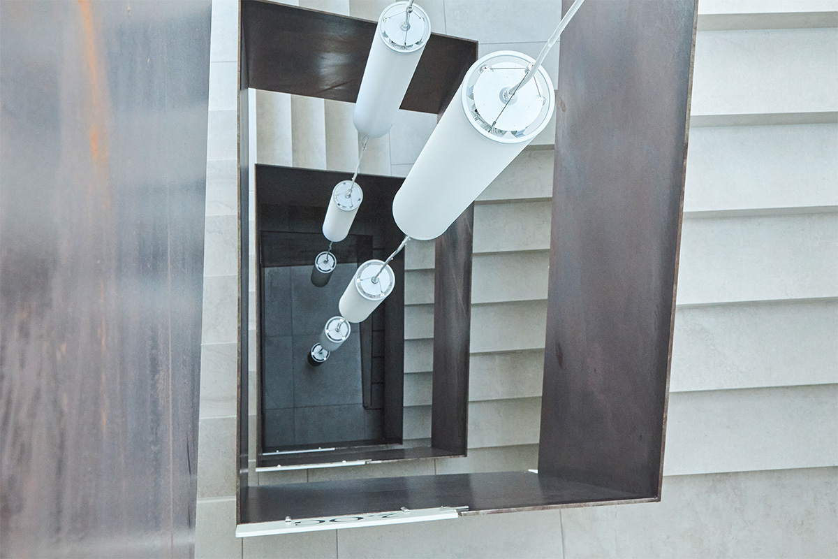 Blick von oben in ein modernes Treppenhaus mit mehreren Etagen und hängenden Deckenleuchten.