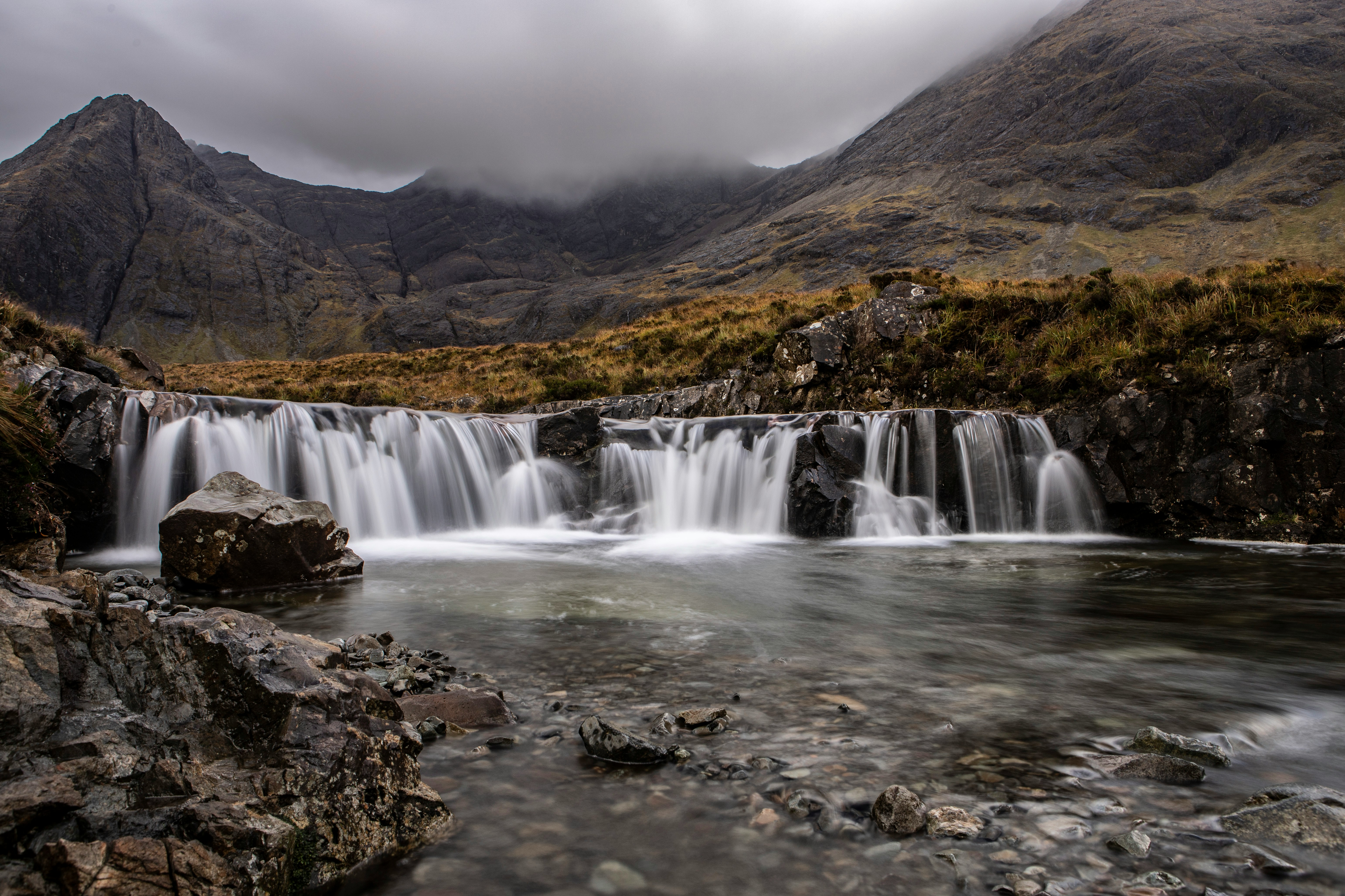 Ein kleiner Wasserfall flie&szlig;t in einen klaren Bach, umgeben von Felsen und einer rauen Berglandschaft unter tiefh&auml;ngenden Wolken. Foto von Angelo Casto auf Unsplash